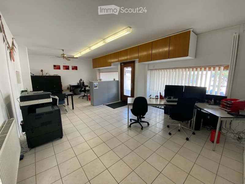 Office space with tiled floor, white walls, ceiling lights, and a large window with blinds. Includes office chairs, tables, a printer, and cabinets.