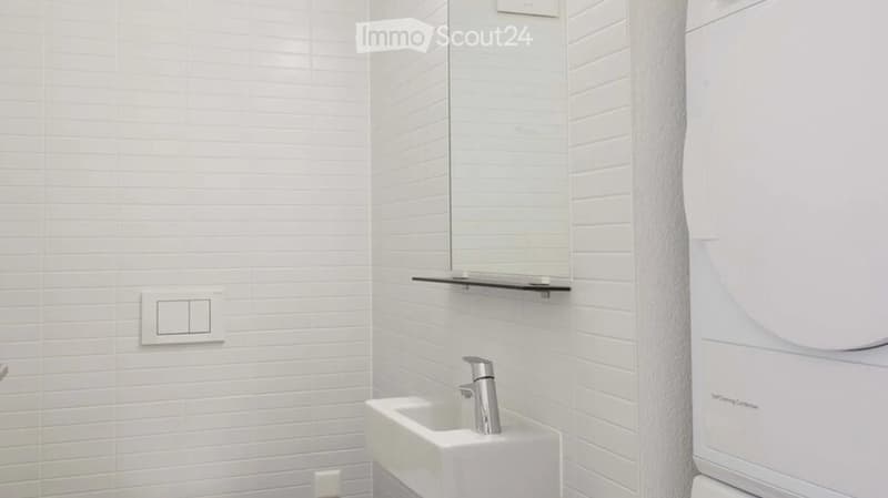 Modern bathroom with white tiled walls, a white sink with a chrome faucet, a mirror above the sink, and a shelf for storage.