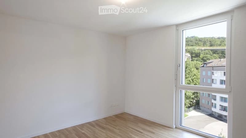 Empty room with light-colored walls and wooden flooring, featuring a large window with a view of greenery and buildings.