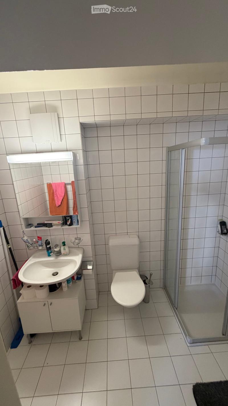 A compact bathroom with white tiled walls and floor, featuring a sink with a cabinet, a toilet, a mirror, and a glass-enclosed shower. A pink towel hangs on a rack, and various toiletries are placed on the sink counter.