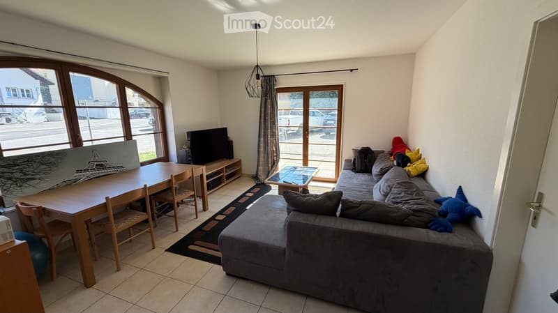 Living room with a grey sectional sofa, wooden dining table with chairs, TV, and a large window with a view of the street.