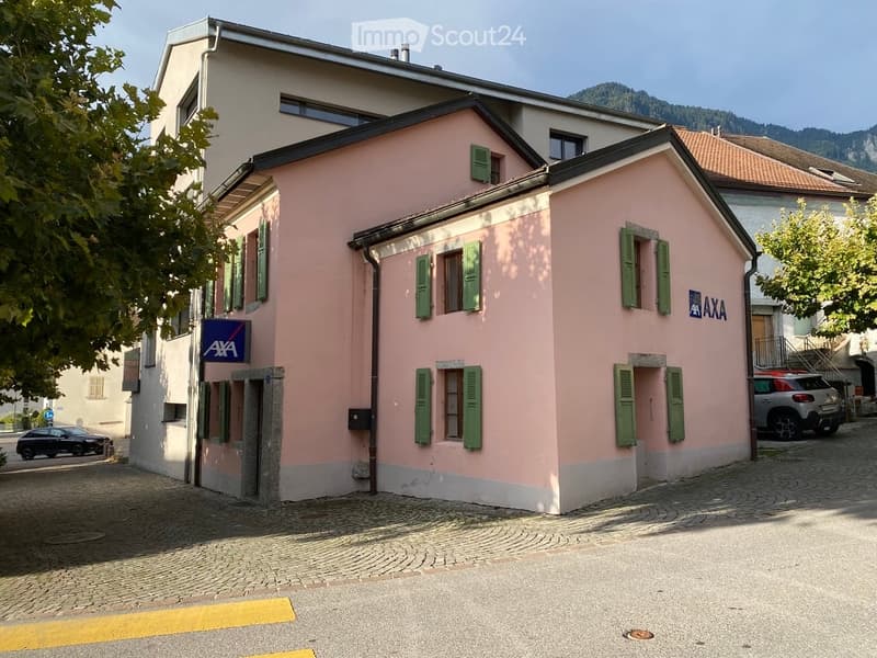 A two-story pink building with green shutters and a sign reading "AXA" on the facade, situated on a cobblestone street with a parked car nearby.