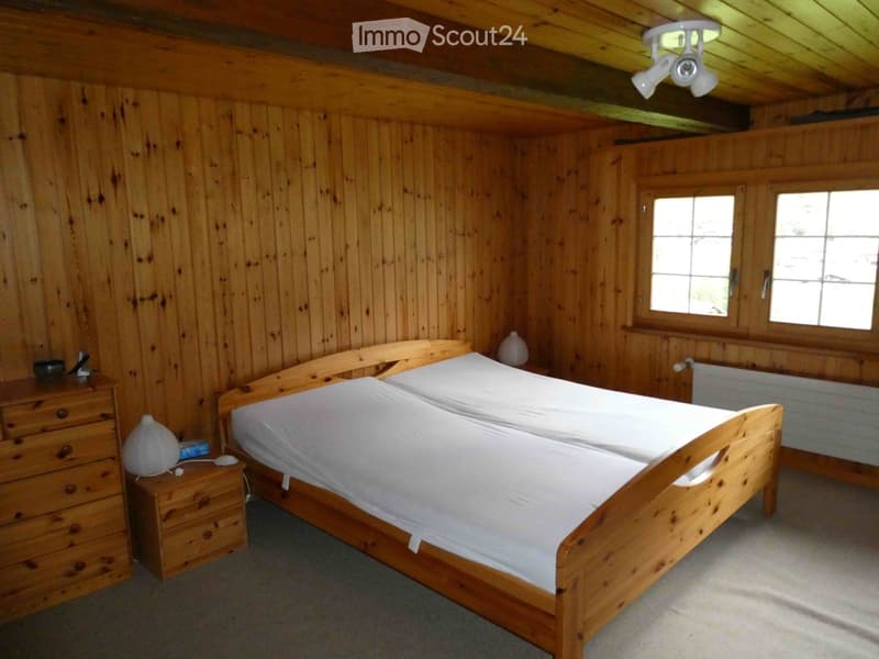 Cozy wooden bedroom with a double bed, nightstand, lamp, and radiator. The room features wooden walls and a slanted wooden ceiling.