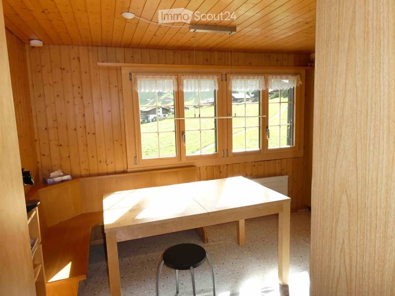 Bright room with wooden walls and ceiling, large windows with white curtains, a wooden table, and a black stool.