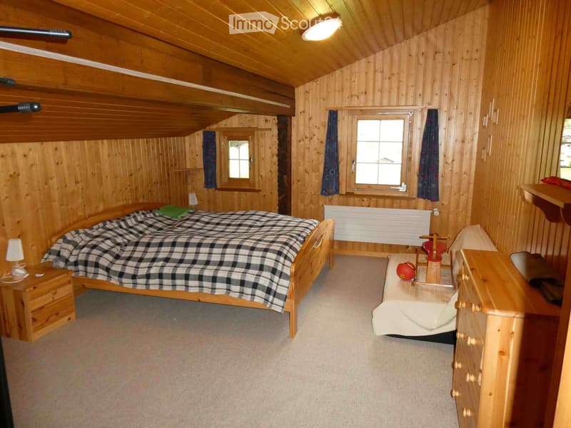 Cozy wooden bedroom with a double bed, nightstand, lamp, and a small chair with a ball on it. The room features wooden walls and ceiling, two windows with curtains, and a radiator.