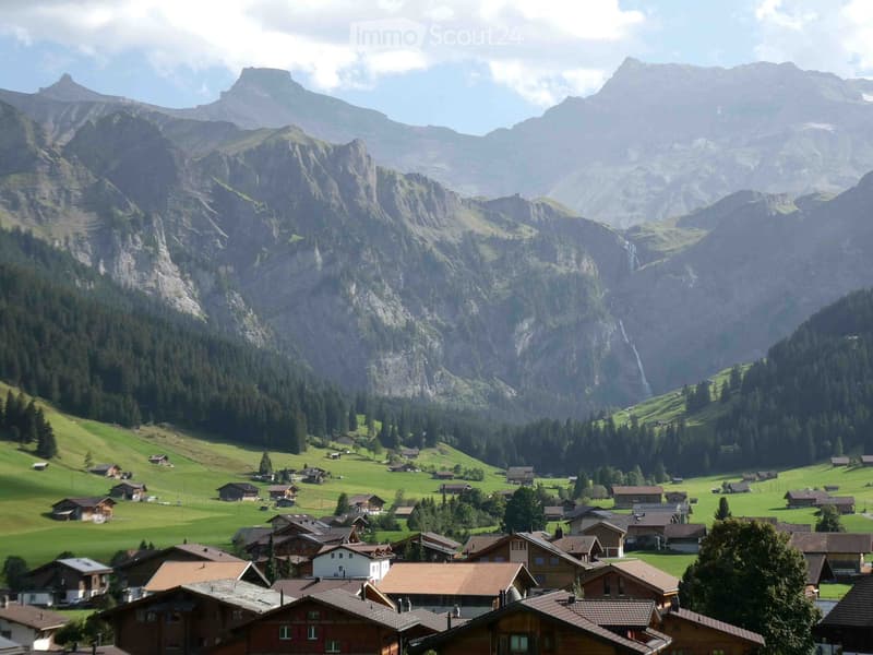 A picturesque village nestled in a lush green valley surrounded by towering mountains. The village features traditional wooden houses with red-tiled roofs scattered across the landscape, with dense forests covering the slopes of the mountains.