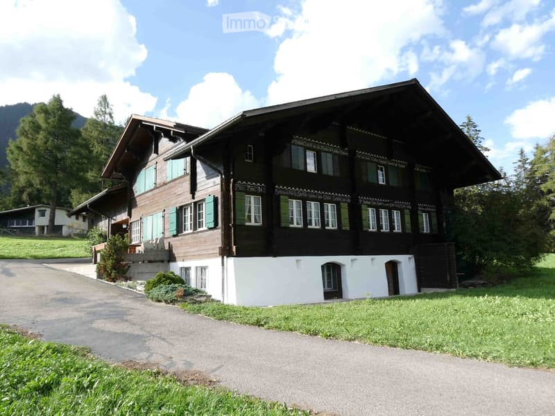 A large, traditional wooden house with a steeply pitched roof, multiple windows, and decorative wooden trim. The house is surrounded by a well-maintained lawn and trees, with a gravel driveway leading to the entrance.