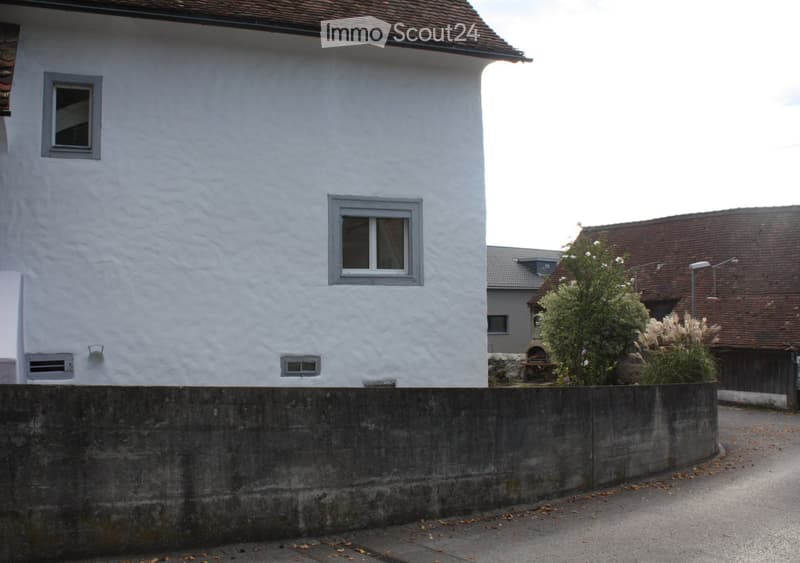 White plastered wall, brown roof, grey concrete fence, two windows, a window on the corner