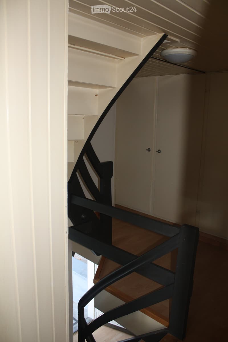 A spiral staircase with a black handrail, wooden steps, and a bright white ceiling light