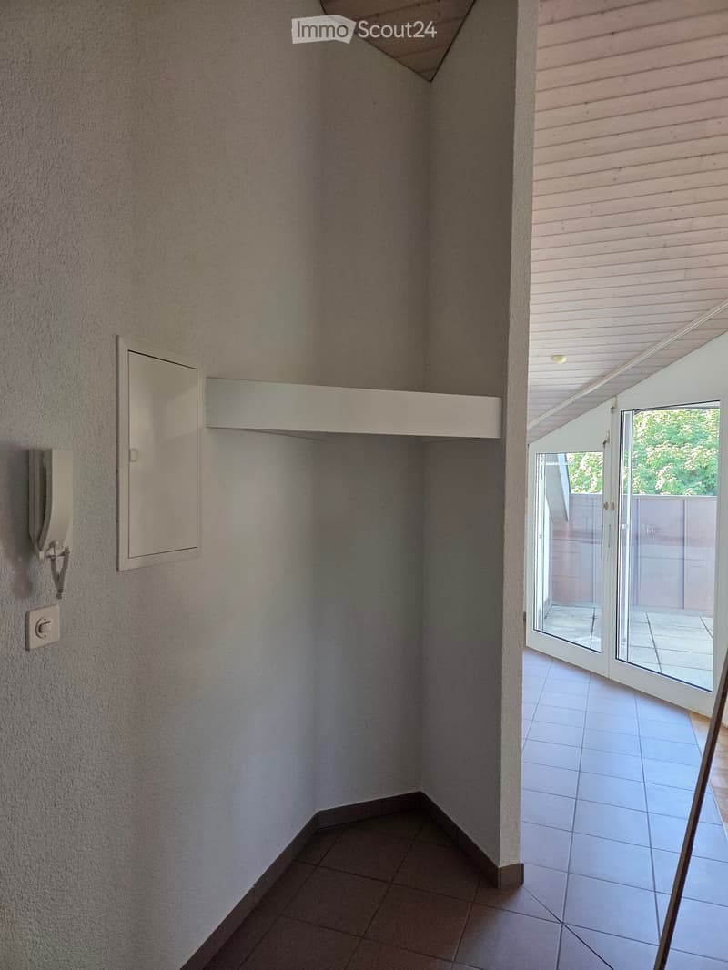 A narrow hallway with tiled flooring, light-colored walls, and an open sliding door leading to a balcony area with outdoor tiles and a view of greenery.
