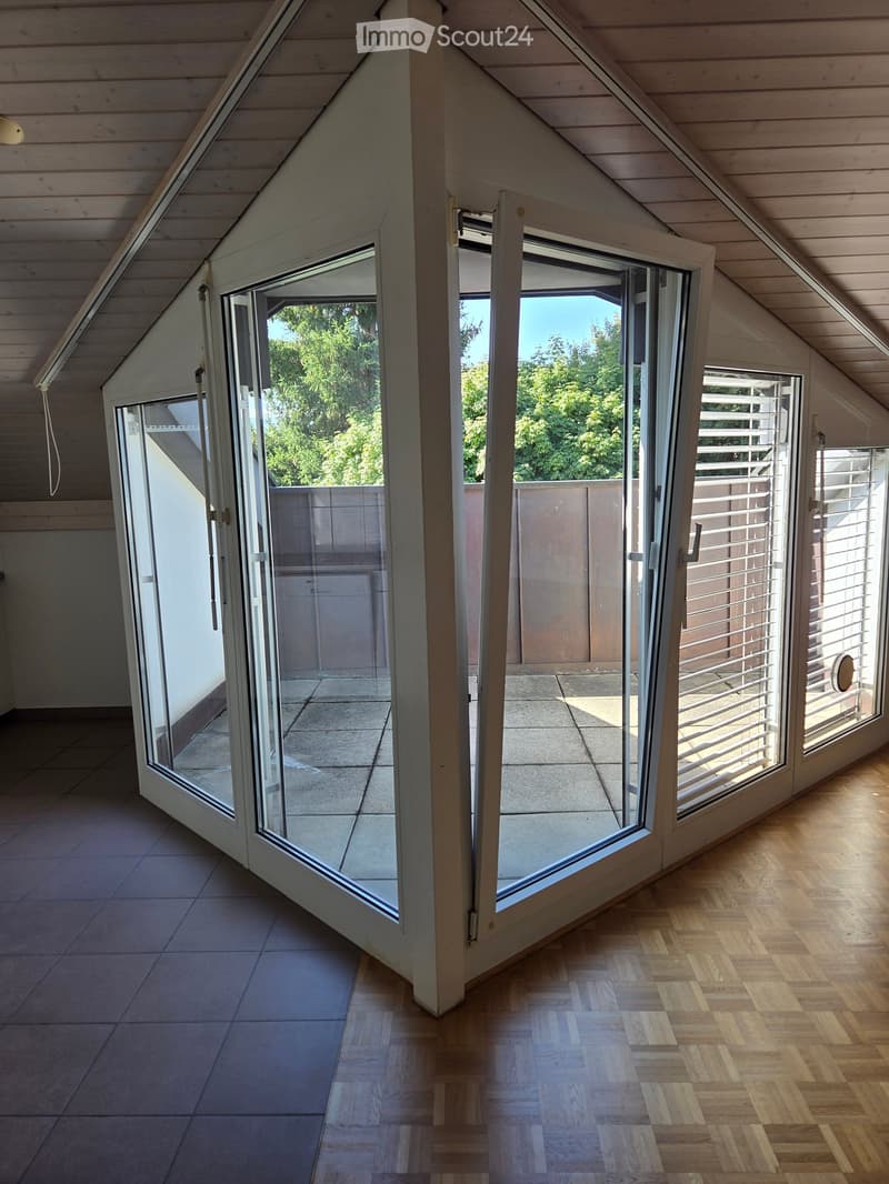 Bright, empty attic room with wooden floors, large sliding glass doors leading to a balcony, and a view of greenery outside.