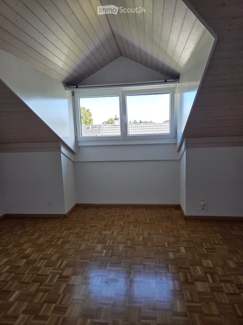 Empty attic room with wooden herringbone flooring, white walls, and skylight windows offering a view of rooftops and trees.