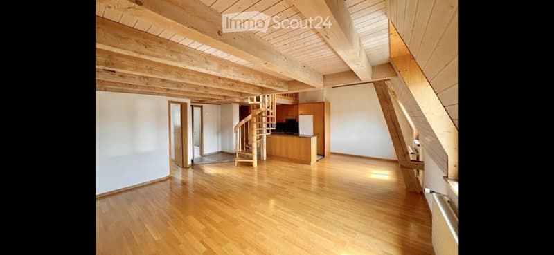 Open, spacious room with wooden flooring and exposed wooden beams on the ceiling. Features a spiral staircase leading to an upper level and a small kitchen area with cabinets and appliances.