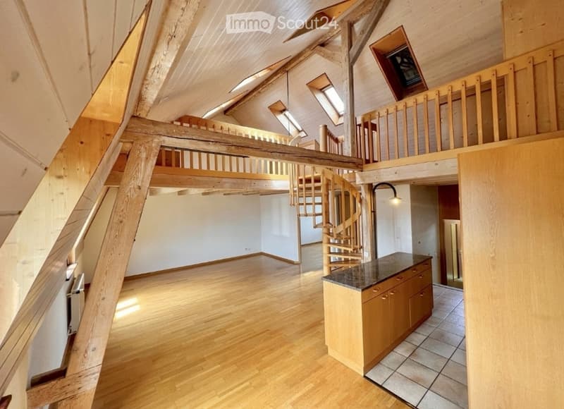 Open-plan attic space with wooden beams, exposed wooden floor, spiral staircase leading to a loft area, and a small kitchen counter with cabinets.