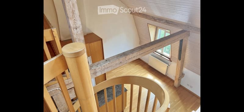 Spiral staircase with wooden railings and polished wooden floors, leading to a room with large windows and white walls.