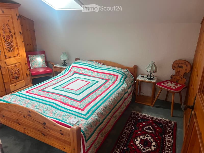 Cozy bedroom with wooden bed, colorful quilt, nightstands with lamps, wooden wardrobe, and a small seating area with a chair and stool.