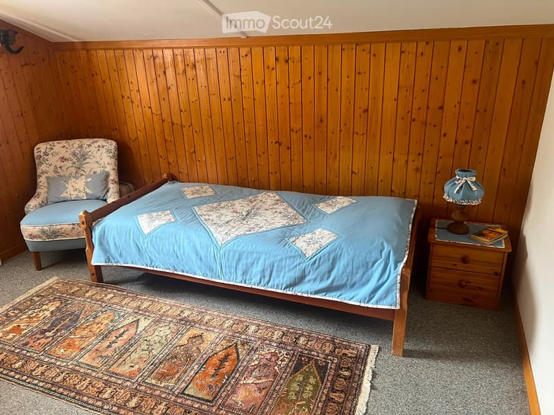 Cozy bedroom with wooden walls, a single bed with blue bedding, a patterned rug, a wooden nightstand with a table lamp, and a floral-patterned armchair.