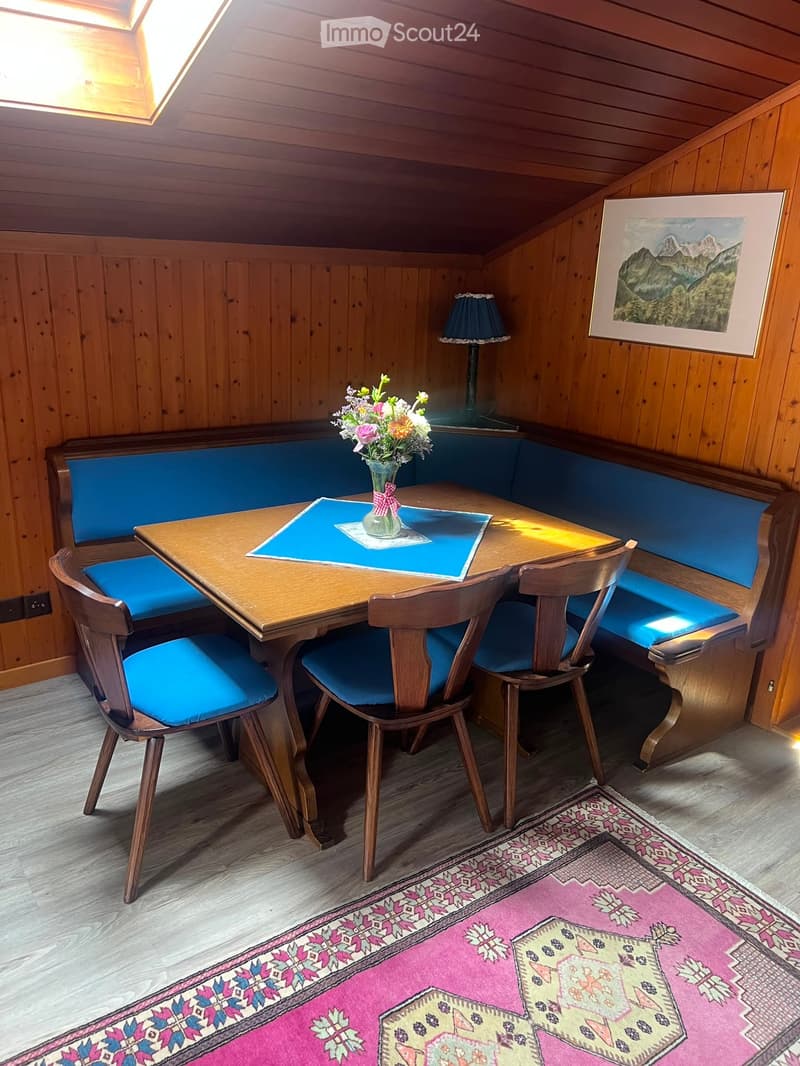 Cozy wooden-paneled room with a round wooden table, four chairs, a built-in bench, a vase with flowers, a lamp, and a framed picture on the wall.