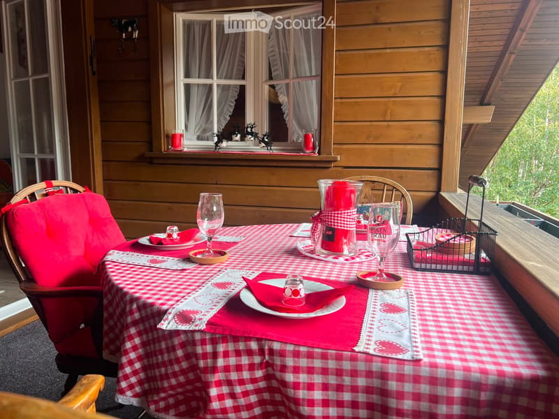 A cozy balcony with a red and white checkered tablecloth, set for two with wine glasses, plates, and candles, overlooking a green landscape.