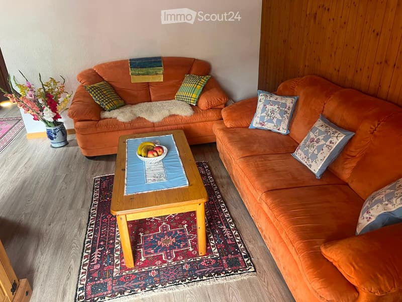 Cozy living room with an orange sectional sofa, wooden coffee table, decorative pillows, and a vase with flowers on the floor.