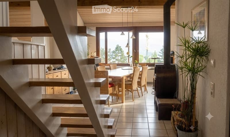 A modern, open-plan living and dining area with a wooden staircase leading upwards, a dining table with chairs, a fireplace, and large windows offering a scenic view.