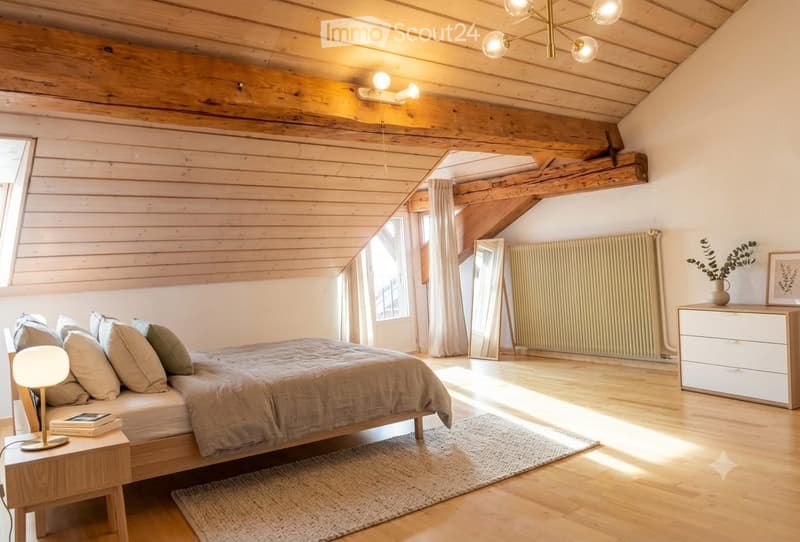 Cozy attic bedroom with wooden beams, slanted ceiling, and large windows. Features a bed with pillows and a duvet, a nightstand with a lamp, a mirror, a radiator, a drawer unit, and a small plant on the dresser.