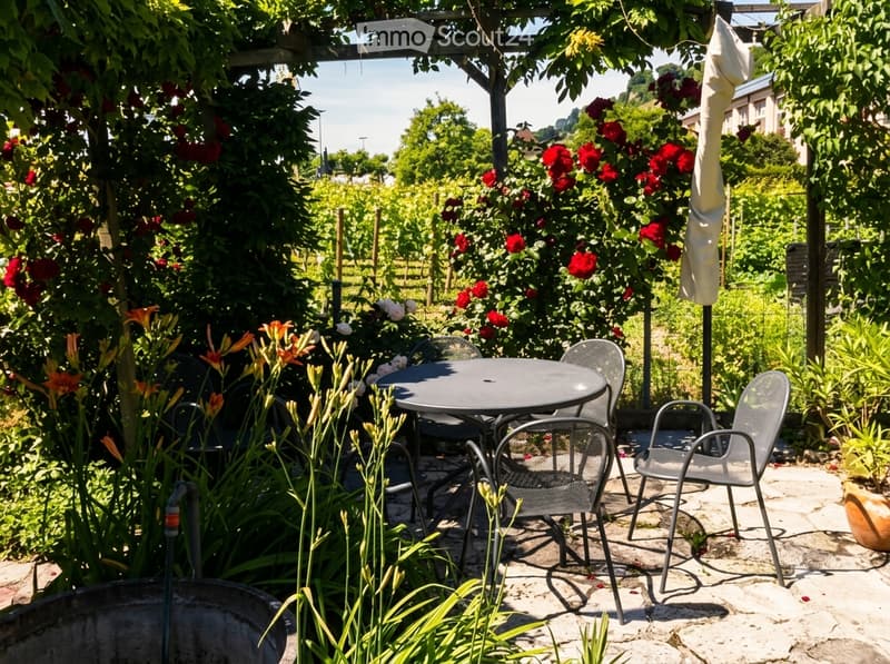 A well-maintained garden with a patio area featuring a round metal table and four matching chairs, surrounded by vibrant flowers and lush greenery.