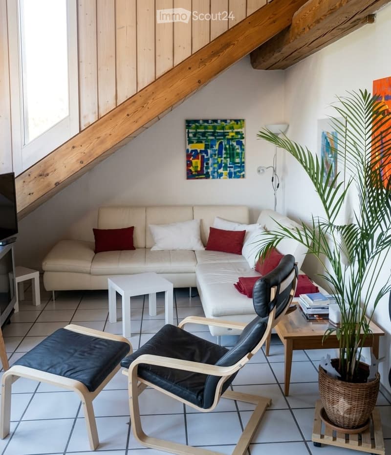 Modern living room with a white sectional sofa, red throw pillows, a small white coffee table, a wooden side table, a black leather chair, and a potted plant. The room features exposed wooden beams and a slanted ceiling.