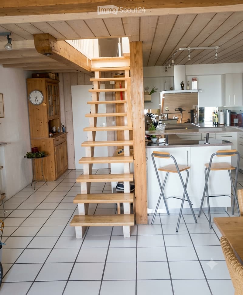 The image shows a modern, open-plan living and kitchen area with a wooden staircase leading to an upper level. The kitchen features a white cabinet, a kitchen island with bar stools, and various appliances. A wooden cabinet with glass doors and a clock is