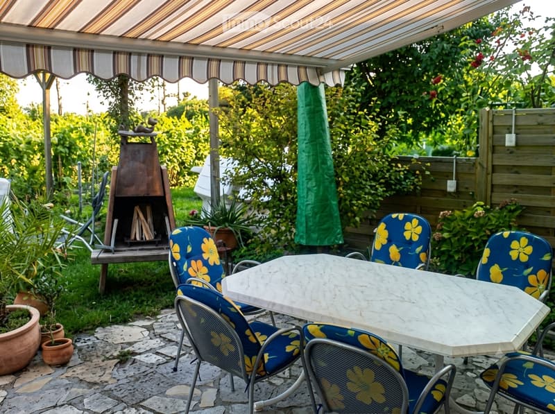 Garden area with a covered patio, a marble table, four blue chairs with yellow floral cushions, a barbecue, potted plants, and a green umbrella.