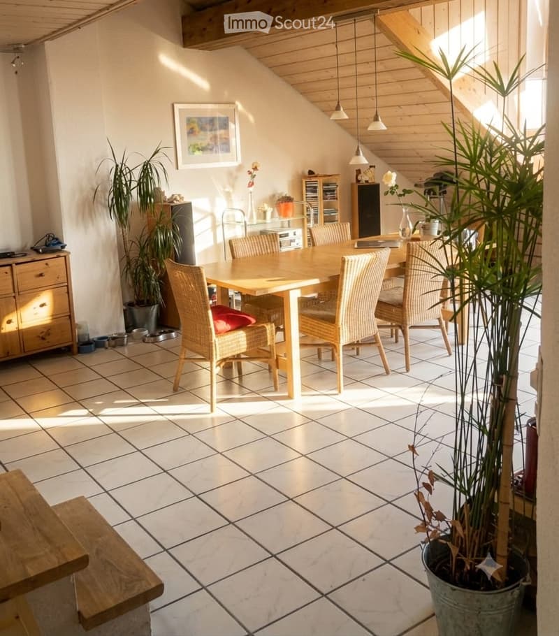 Bright dining area with a wooden dining table surrounded by wicker chairs, a wooden bench, and potted plants. The space features tiled flooring and wooden ceiling beams.