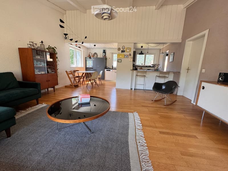 Open-plan living and dining area with wooden flooring, a green sofa, a round coffee table, a dining table with chairs, a kitchen island, and various decorative items.