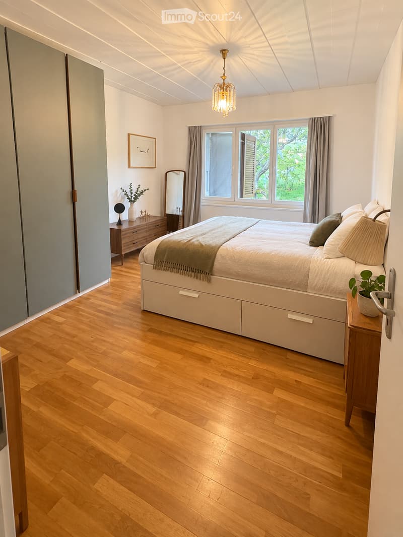 Modern bedroom with wooden flooring, large bed with drawers, nightstand, mirror, dresser, and decorative items.