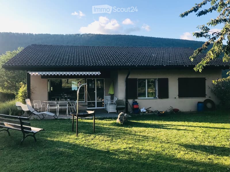 A well-maintained garden area with a wooden bench, several white chairs, a table, a swing, and a fire pit. The house in the background has a tiled roof and large windows, with a scenic mountain view.