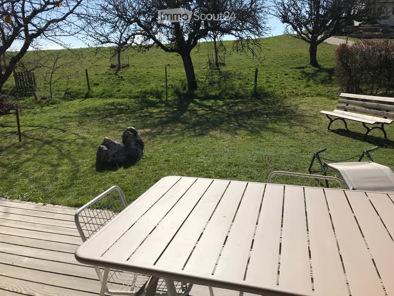 Outdoor garden area with a wooden deck, white plastic table, and chairs, surrounded by grassy lawn and trees.