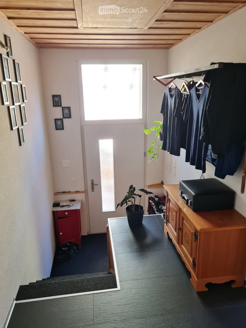 Hallway with wooden ceiling, white walls, a door leading outside, coat rack with hanging clothes, small table with potted plant, and a staircase leading upwards.