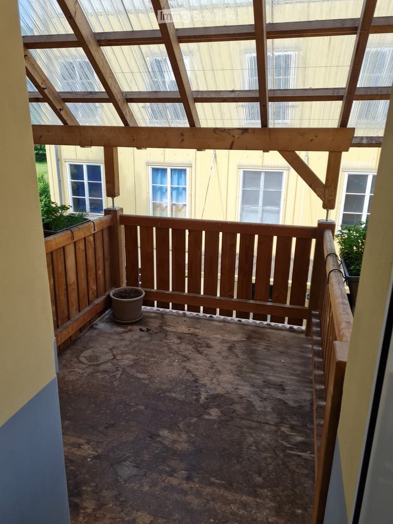 Wooden balcony with a railing, a small potted plant, and a view of a yellow building with windows.