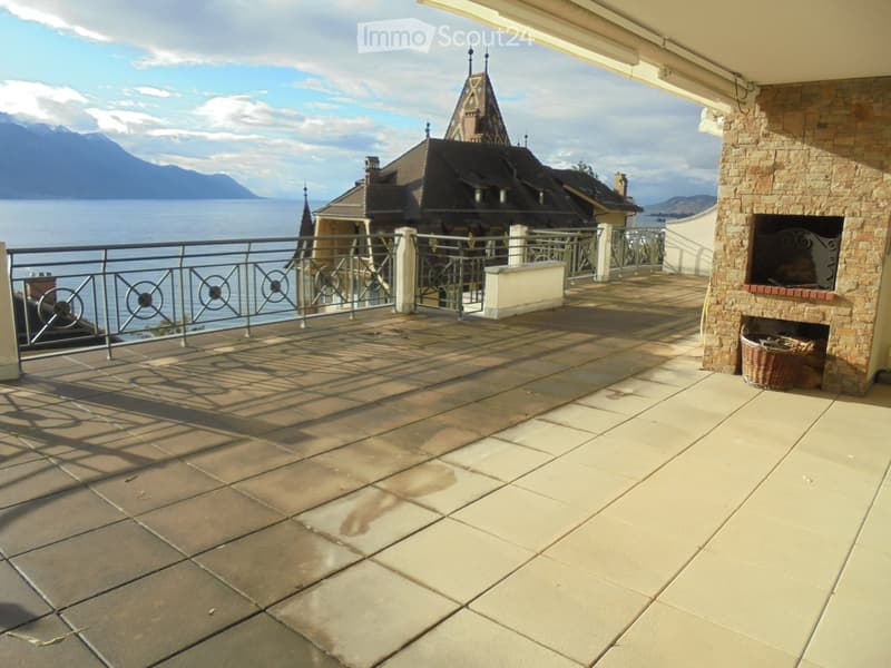 Balcony with stone tiles, metal railing, and a view of a lake and mountains. Features a brick fireplace and a small basket.