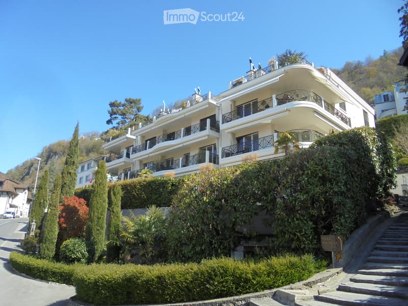 A large, multi-story villa with white walls, multiple balconies, and decorative railings, surrounded by lush greenery and trees.