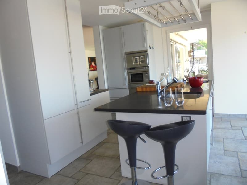 Modern kitchen with white cabinets, black countertop, two black bar stools, and a built-in oven. The island features a sink with a modern faucet, and there are glass cups and a red bowl on the counter.