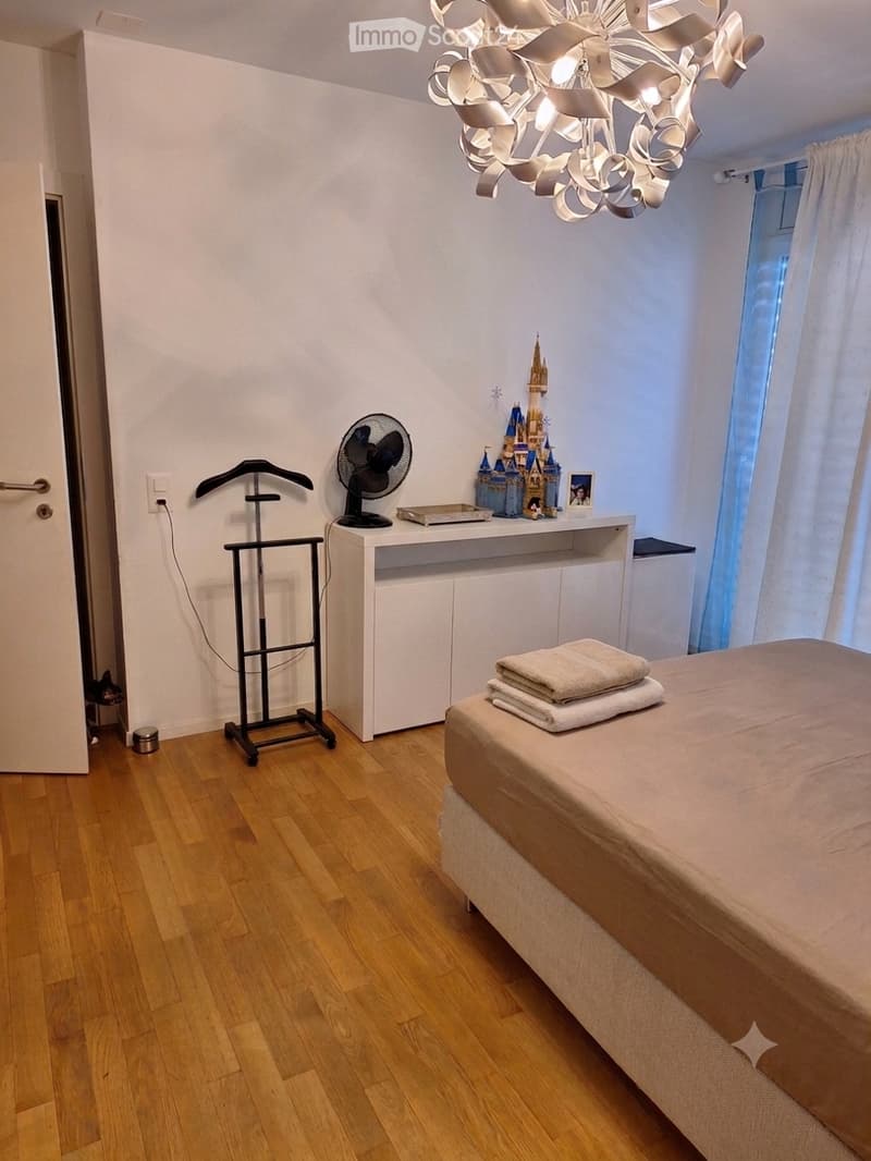 A well-lit bedroom with wooden flooring, a bed with beige bedding, a small table with a fan and decorative items, and a wardrobe. The room features large windows with white curtains and a modern chandelier.