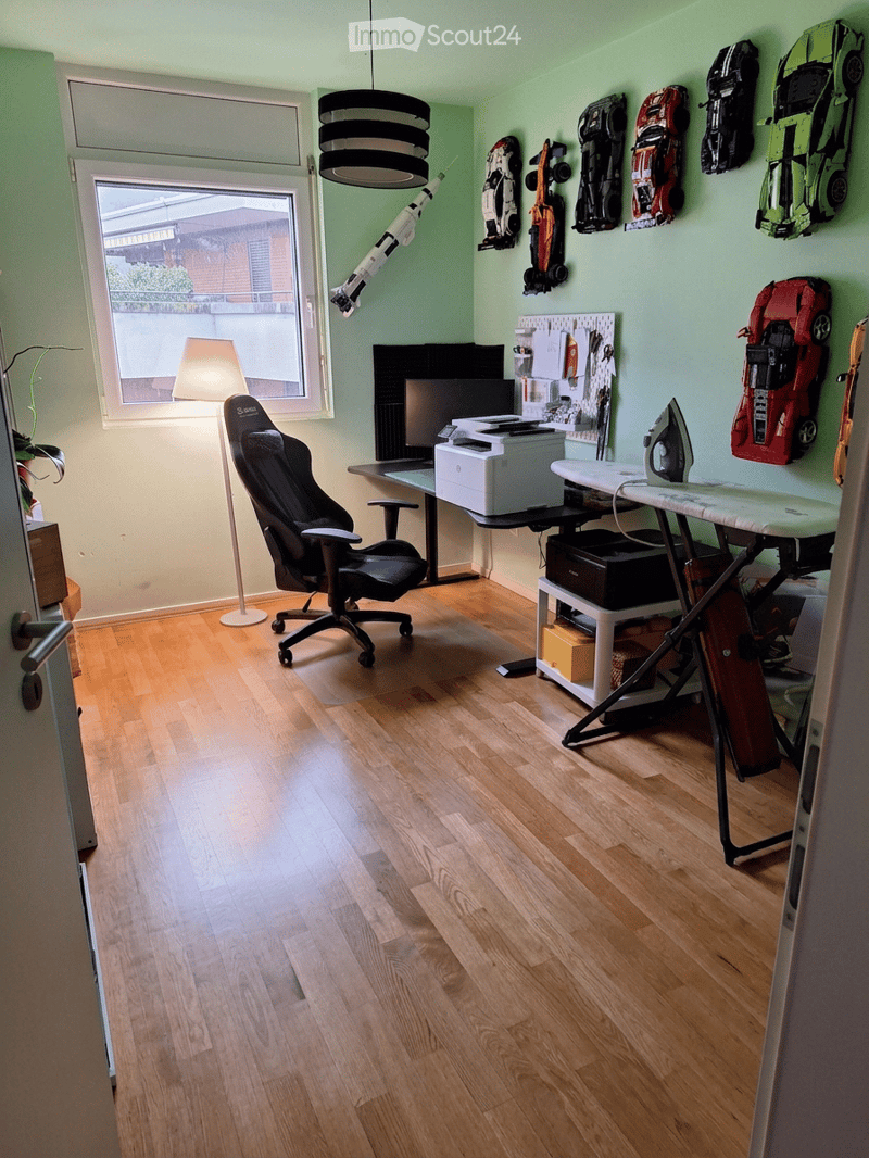 A well-lit room with wooden flooring, a large window, a black office chair, a desk with a computer screen and printer, an ironing board, and several toy cars on the wall.