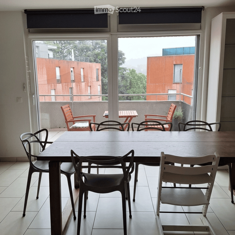 A modern dining area with a large wooden table and several black chairs, overlooking a balcony with outdoor seating and a view of neighboring buildings.
