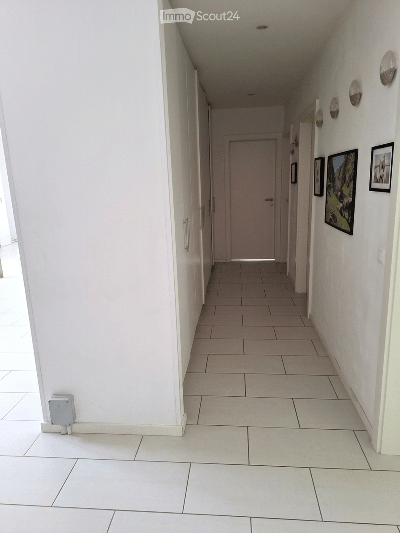 A clean, well-lit hallway with white tiled flooring and walls, featuring several doors and framed artwork on the right side.