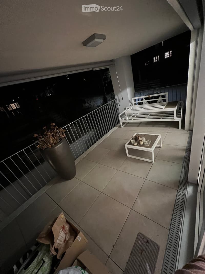 Balcony with tiled floor, white table and chairs, flower pot, and railing overlooking the night view.