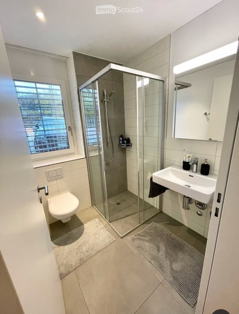 Modern bathroom with a glass-enclosed shower, toilet, sink, and mirror. Features tiled walls and a window with blinds.