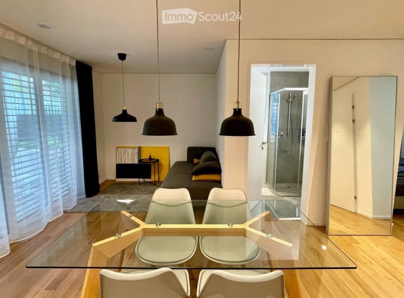 Modern living and dining area with a glass dining table, white chairs, a sofa, and a small coffee table. The room features wooden flooring, large windows with blinds, and pendant lights. A shower is visible in the adjacent bathroom.