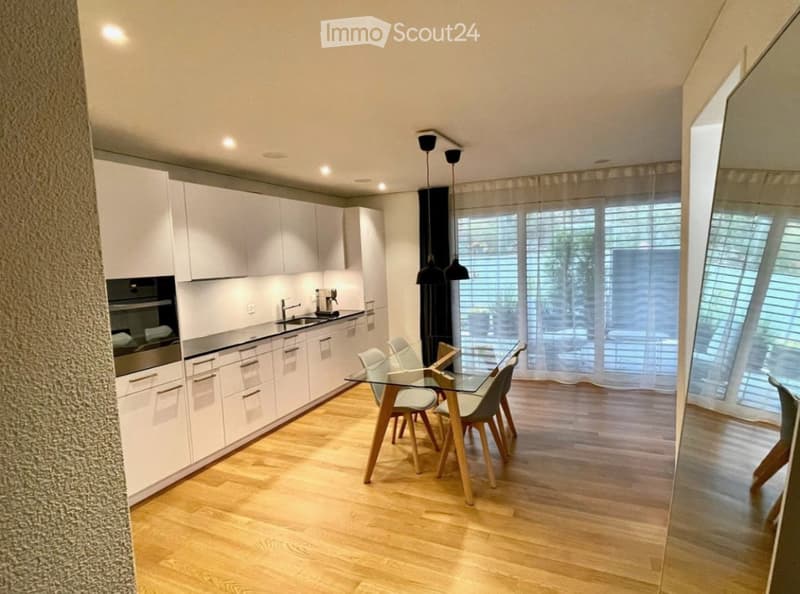 Modern kitchen with white cabinetry, stainless steel appliances, a glass dining table with wooden chairs, and large windows with blinds.