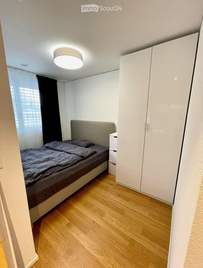 Modern bedroom with a single bed, grey bedding, white wardrobe, and a nightstand. The room features wooden flooring and a large window with black curtains.