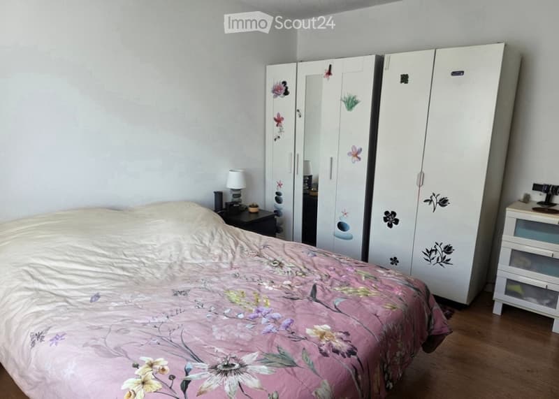 Bedroom with a double bed covered in a pink floral duvet, a large white wardrobe with floral decals, a nightstand with a lamp, and wooden flooring.